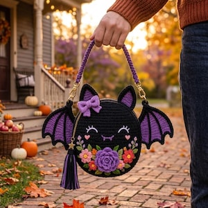 May include: A round, black crocheted bag shaped like a bat with purple inner ears and wings. The front features a purple bow, embroidered closed eyes, a small mouth with fangs, and a cluster of colorful crocheted flowers. Includes a tassel.