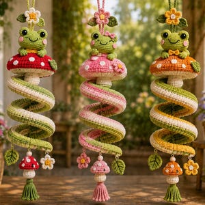 May include: Three hanging crochet decorations featuring green frogs sitting on spotted mushrooms. Each piece has a spiral body in green, pink, or yellow, accented with small flowers, leaves, and a miniature mushroom at the bottom with a tassel.