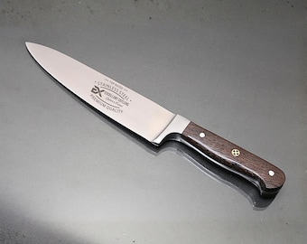 May include: A stainless steel chef knife with a dark brown wood handle. The blade features the text TOP RATED STAINLESS STEEL EX EXCELLENT CUTLERS PREMIUM QUALITY. The handle is secured with two silver rivets and a central decorative brass pin.