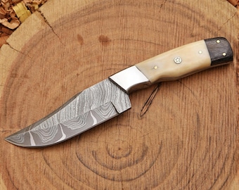 May include: A fixed-blade knife featuring a patterned Damascus steel blade with a wavy, layered aesthetic. The handle is constructed from a cream-colored material accented with a dark wood end cap, secured by three small metal pins and a central rivet.