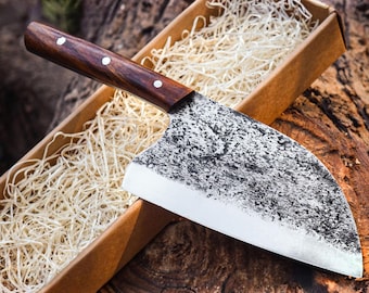 May include: A cleaver with a dark brown wooden handle secured by three silver rivets and a wide, curved blade featuring a textured, hammered metal finish. The knife rests inside a rectangular cardboard box filled with light-colored wood shavings.