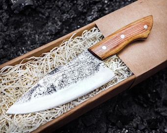 May include: A fixed-blade knife with a wide, textured, dark-finished steel blade and a light brown wooden handle secured by three silver-colored rivets. The knife is nestled inside a rectangular cardboard box filled with light-colored wood shavings.