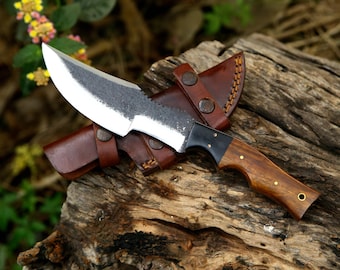 May include: A fixed-blade knife with a polished steel edge and a dark, textured spine. The handle features a two-tone design with a black bolster and a polished wooden grip secured by brass pins. It is paired with a brown leather sheath.