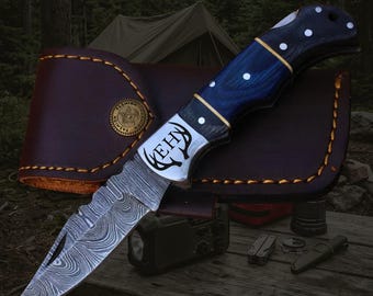 May include: A folding pocket knife with a Damascus steel blade featuring a swirling pattern and a bolster engraved with the letters EHY and an antler design. The handle is dark blue wood with white dots and a brown leather sheath with gold stitching.