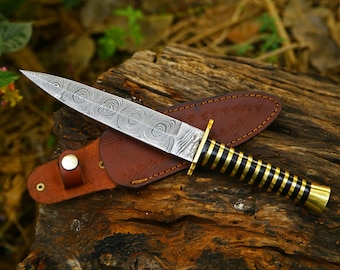 May include: A fixed-blade knife with a Damascus steel blade featuring a swirling pattern. The handle consists of alternating black and brass rings, and the knife includes a brown leather sheath with a metal snap closure.