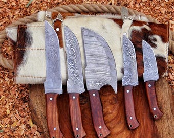May include: A set of five Damascus steel knives with dark brown wooden handles and silver rivets. The blades feature a distinct wavy pattern. The knives are arranged in a row, resting against a white and brown cowhide roll with rope ties.