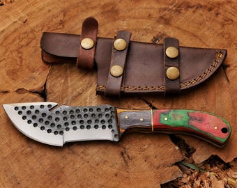May include: A fixed-blade knife with a dark, hammered metal blade featuring a row of circular indentations and a gut hook. The handle is constructed from layered wood in red, green, and dark brown tones. A brown leather sheath with brass snaps.