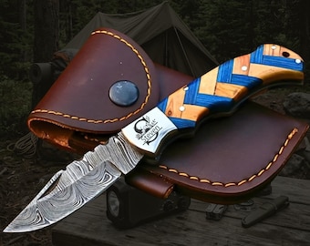 May include: A folding pocket knife with a Damascus steel blade and a handle featuring a blue and natural wood herringbone pattern. The metal bolster is engraved with the name Steven. The knife rests on a brown leather sheath with yellow stitching.