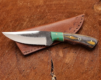 May include: A fixed-blade knife with a polished steel blade, a bright green bolster, and a dark wood handle featuring yellow grain accents and brass pins. The knife rests on a brown leather sheath with decorative stitching and embossed details.