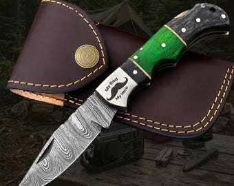 May include: A folding pocket knife with a Damascus steel blade and a handle featuring green and black wood segments. The metal bolster is engraved with a mustache icon and the text My Dad My Hero. It is paired with a brown leather sheath.