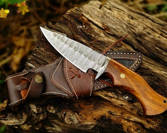 May include: A fixed-blade knife featuring a patterned Damascus steel blade and a polished wooden handle with a brass pin. The knife is accompanied by a brown leather sheath with visible stitching, a metal snap button, and a leather cord.