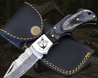 May include: A folding knife with a Damascus steel blade and a dark wood handle featuring brass rivets. The bolster is engraved with an anchor icon, the text "HENRY WILSON", and "2ND SEPTEMBER 2026". It rests on a black leather sheath with yellow stitching.