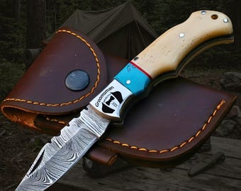 May include: A folding pocket knife with a Damascus steel blade, a cream-colored handle, and a blue bolster. The bolster features a graphic of a tuxedo and the text Groomsman. The knife rests on a brown leather sheath with orange stitching.