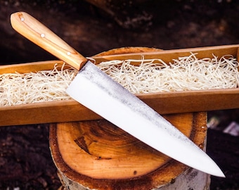 May include: A kitchen knife with a polished silver metal blade and a light brown wooden handle secured by three small metal rivets. The knife is positioned diagonally across a rectangular brown cardboard box filled with light-colored wood shavings.