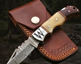 May include: A folding pocket knife with a Damascus steel blade featuring a wavy pattern, a cream-colored handle, and a dark wood bolster. The metal bolster is engraved with the letter W and the name WILSON. It rests on a brown leather sheath.