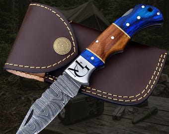 May include: A folding pocket knife with a Damascus steel blade featuring a wavy pattern and a handle made of brown wood and blue resin. The bolster is engraved with the letters MG. The knife is displayed with a brown leather sheath with stitching.