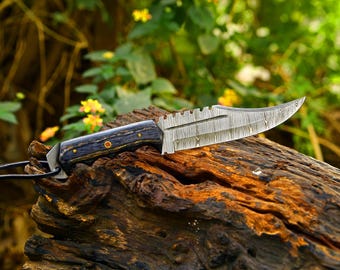 May include: A fixed-blade knife featuring a Damascus steel blade with a serrated spine and a dark, textured wooden handle secured with two brass pins. A black leather lanyard is attached to the pommel, which has a metallic bolster.