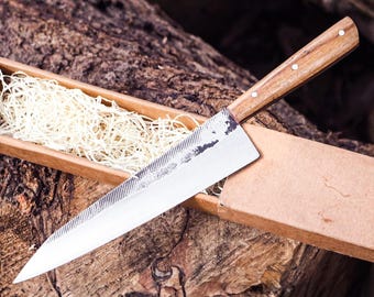 May include: A fixed-blade knife with a polished steel blade featuring a textured, dark metal spine and a wooden handle secured with three metal rivets. The knife is presented in a rectangular cardboard box filled with light-colored wood shavings.