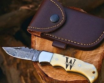 May include: A folding pocket knife with a patterned Damascus steel blade and a cream-colored handle featuring the engraved letter W and the name WILSON. The knife is accompanied by a dark brown leather sheath with visible yellow stitching.
