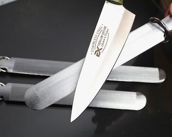 May include: A stainless steel kitchen knife with a pointed blade and the text "STAINLESS STEEL EXCELLENT CUTLERS Quicker & Fastest" printed on the side, positioned over three flat, rectangular metal sharpening stones with attached metal rings.