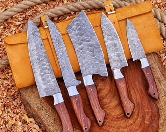 May include: A set of five kitchen knives with Damascus steel blades featuring a wavy pattern and dark brown wooden handles with metal rivets, arranged on a wooden surface in front of a tan leather knife roll with a buckle closure.