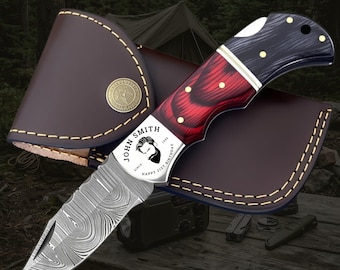 May include: A folding pocket knife with a Damascus steel blade and a two-tone red and black wood handle. The bolster features the text "JOHN SMITH" and "HAPPY 21ST BIRTHDAY" with a bearded face icon. It is paired with a brown leather sheath.