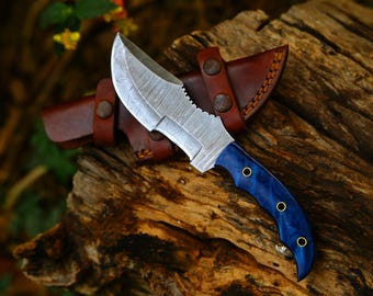 May include: A fixed-blade knife featuring a patterned Damascus steel blade with a serrated spine section and a polished blue resin handle secured by three brass rivets, accompanied by a brown leather sheath with two metal snap fasteners.