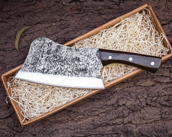 May include: A meat cleaver with a wide, textured, dark gray hammered metal blade and a smooth, dark brown wooden handle secured with three silver-colored rivets, resting inside a rectangular cardboard box filled with light-colored wood shavings.