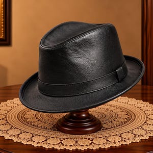 Puede incluir: Un sombrero fedora de cuero negro que presenta una corona estructurada con un pliegue central y una banda de cuero negro a juego que rodea la base. El sombrero tiene un ala curva y una superficie lisa y texturizada, sobre un soporte de madera.