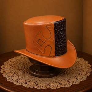 Può includere: Un cappello a cilindro strutturato realizzato in pelle liscia color cuoio, caratterizzato da un prominente motivo decorativo ritagliato sul lato e da un pannello a contrasto marrone scuro con texture goffrata effetto coccodrillo.