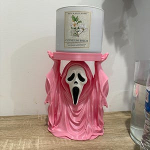 Puede incluir: Un portavelas rosa impreso en 3D con forma de figura fantasma con máscara blanca, que sostiene una vela blanca Bath & Body Works Clothesline Breeze. La etiqueta de la vela muestra flores blancas y hojas verdes con el texto Bath & Body Works.