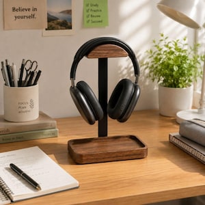 May include: A black over-ear headphone set resting on a vertical stand featuring a dark brown wooden base and a matching wooden top support piece. The stand has a central black metal post connecting the base to the top headphone rest.