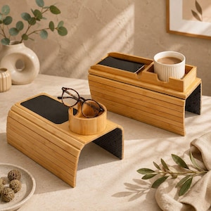 May include: Two bamboo sofa armrest trays with flexible slatted designs. One tray features a black non-slip surface and a circular holder containing brown-rimmed glasses. The other tray holds a smartphone and a white ribbed ceramic mug.
