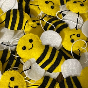 May include: A collection of felt bee decorations with black and yellow stripes, white wings, and black bead eyes. The bees have smiling faces and are arranged in a pile.