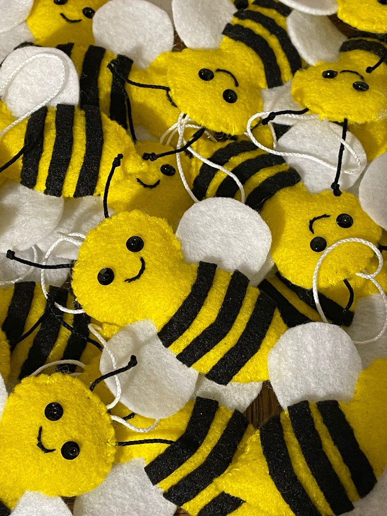 May include: A collection of felt bee decorations with black and yellow stripes, white wings, and black bead eyes. The bees have smiling faces and are arranged in a pile.