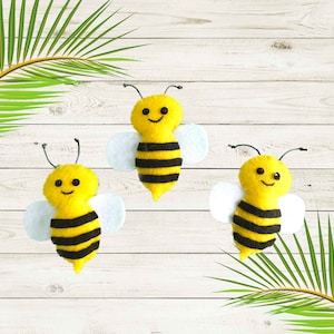 May include: Three felt bee decorations with black and yellow stripes and white wings. The bees have smiling faces and are arranged on a light wood background.