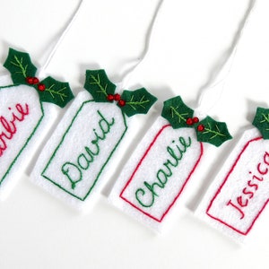 May include: Four white felt Christmas gift tags with green holly leaves and red berries. The tags are embroidered with the names Barbie, David, Charlie, and Jessica.