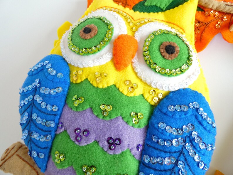 May include: A colorful felt owl with rainbow-colored feathers and blue wings. The owl has large green eyes with white and green sequins around them. The owl is decorated with sequins and has a brown felt beak.