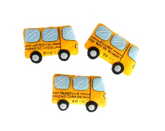 May include: Three yellow felt school bus ornaments with different text on each. The first bus says "Jordyn Preschool 2021". The second bus says "Elijah 1st Day of School 20--". The third bus says "Isabella 2nd Grade 20--".