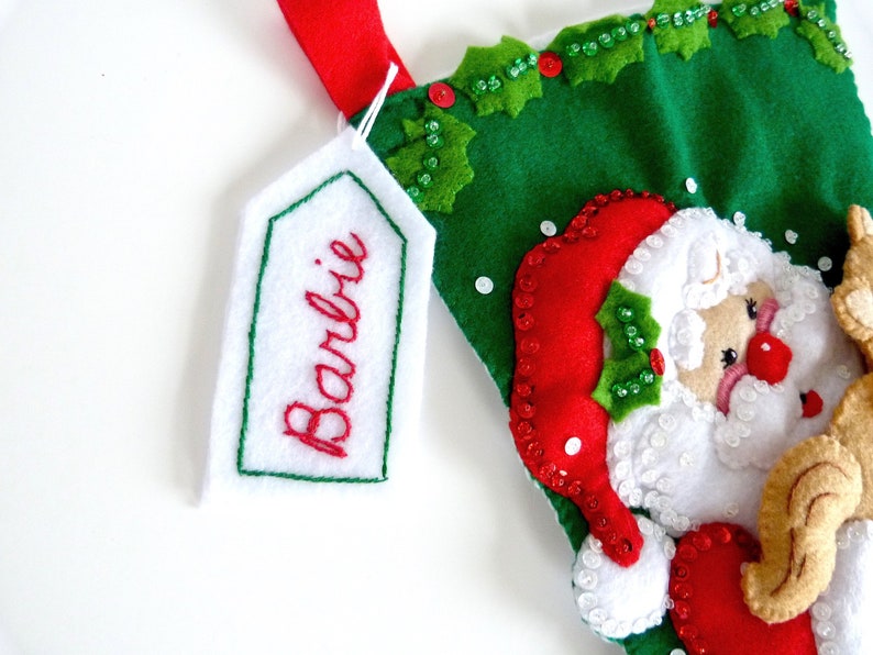 May include: A green felt Christmas stocking with a Santa Claus applique. The stocking has a white felt tag with the name "Barbie" embroidered in red thread.