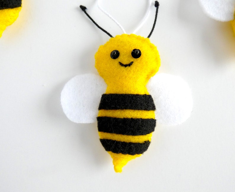 May include: A yellow felt bee with black stripes and white wings. The bee has black eyes and a smiling mouth.