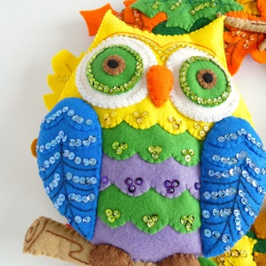 May include: A colorful felt owl with green, yellow, purple, and blue felt. The owl has blue sequin wings and green sequin eyes. The owl is sitting on a brown felt branch.