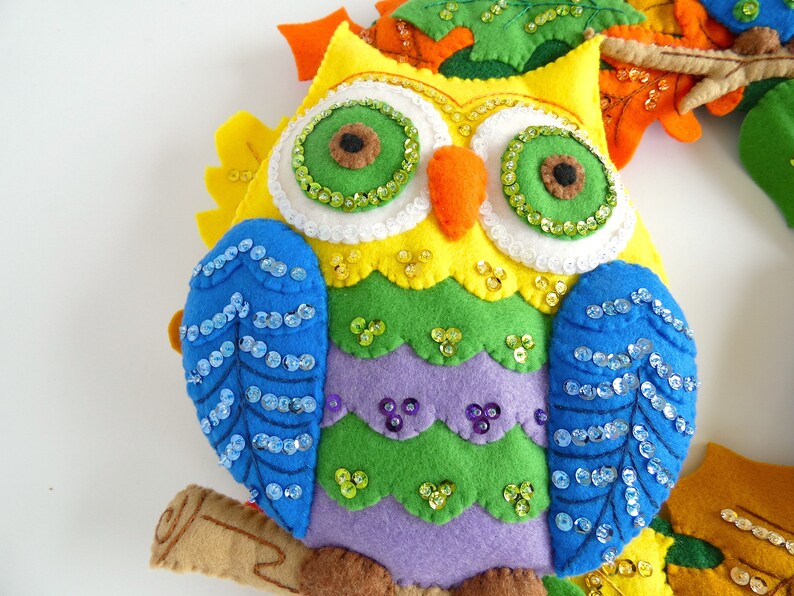 May include: A colorful felt owl with green, yellow, purple, and blue felt. The owl has blue sequin wings and green sequin eyes. The owl is sitting on a brown felt branch.