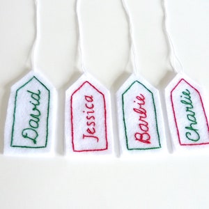 May include: Four white felt gift tags with green and red embroidery. The tags are labeled "David", "Jessica", "Barbie", and "Charlie".