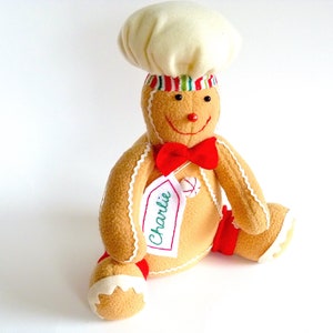 May include: A gingerbread man plush toy wearing a white chef's hat with red and green stripes. The toy has a red bow tie and a name tag that says "Charlie".