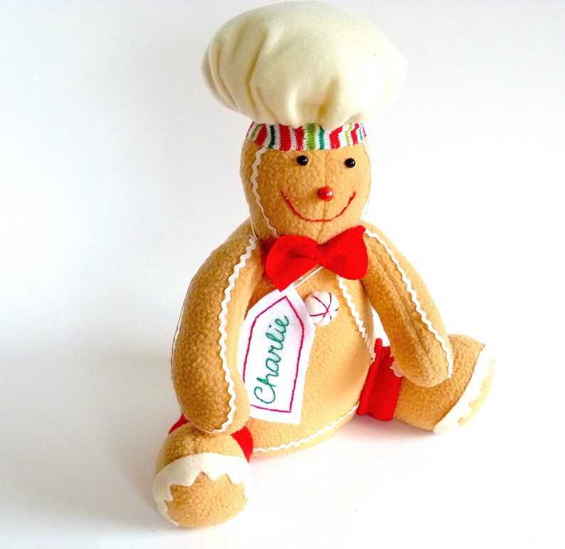 May include: A gingerbread man plush toy wearing a white chef's hat with red and green stripes. The toy has a red bow tie and a name tag that says "Charlie".