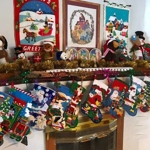 May include: A fireplace mantel decorated for Christmas with stockings, stuffed animals, and festive artwork. The stockings are filled with various items, including a Santa Claus, a snowman, and a train. The artwork includes a winter scene, a nativity scene, and a village scene. The stuffed animals include a dog, a penguin, a gingerbread man, and a snowman.