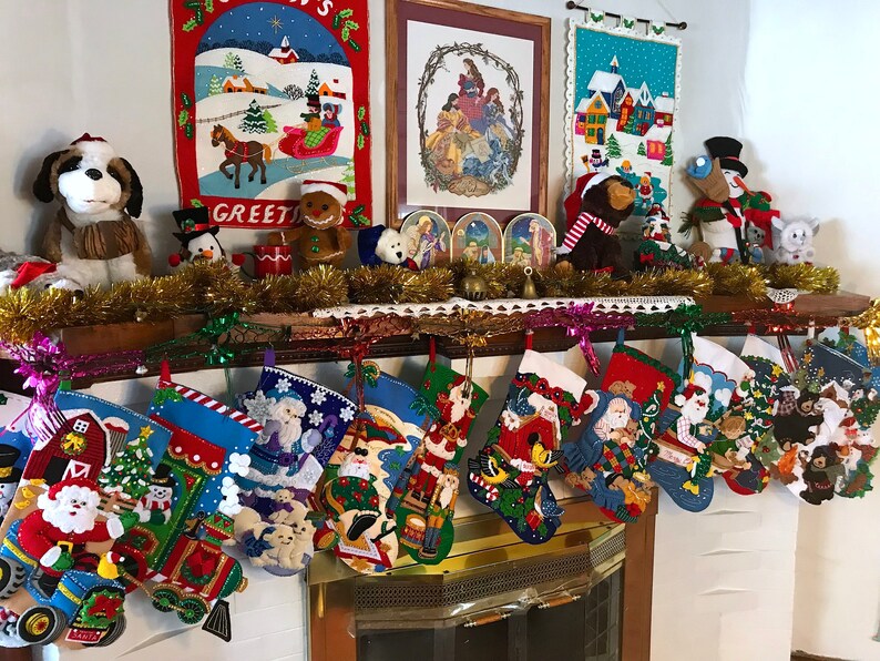 May include: A fireplace mantel decorated for Christmas with stockings, stuffed animals, and festive artwork. The stockings are filled with various items, including a Santa Claus, a snowman, and a train. The artwork includes a winter scene, a nativity scene, and a village scene. The stuffed animals include a dog, a penguin, a gingerbread man, and a snowman.