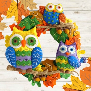 May include: A colorful felt owl wreath with three owls, one yellow, one purple, and one blue, decorated with fall leaves and beads.