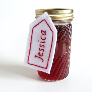 May include: A glass jar with a red liquid inside, topped with a gold lid. A white felt tag with red embroidery reads "Jessica" and is attached to the jar.